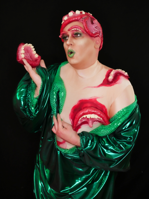Rosalind in a shiny green costume with toothy mouths growing out of her body. Photo credit Good Lucifer.