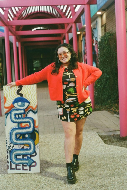 Portrait of the Serena Zam leaning against an artwork in a colorful outdoor walkway.